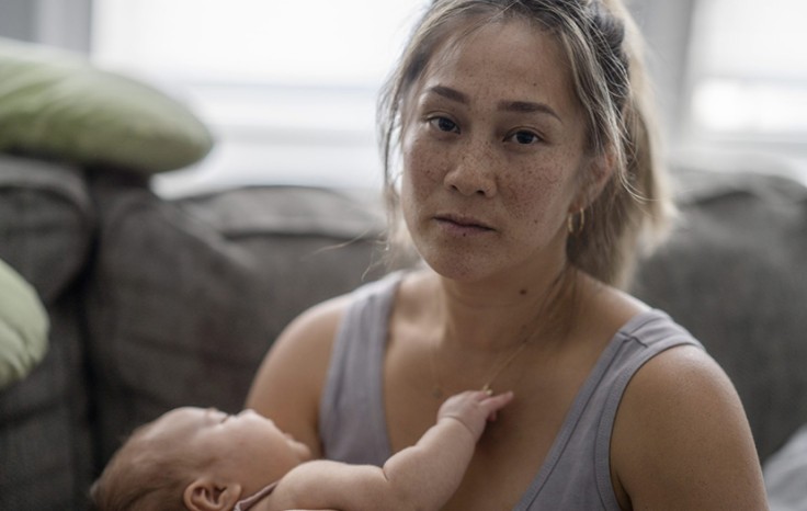 A middle aged mother sitting on her couch holds her baby in her arm with a tired and stressed expression.
